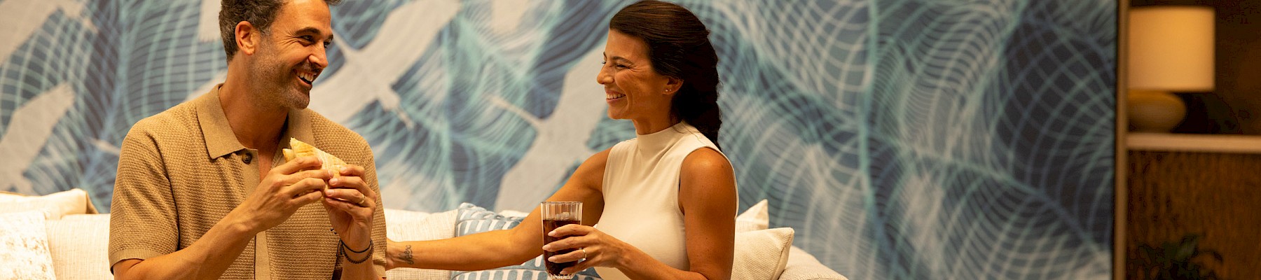 A couple sits on a couch enjoying coffee and snacks, smiling at each other in a stylish, modern lounge with a blue abstract wall behind them.
