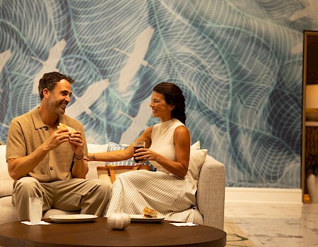 A couple sits on a couch enjoying coffee and snacks, smiling at each other in a stylish, modern lounge with a blue abstract wall behind them.