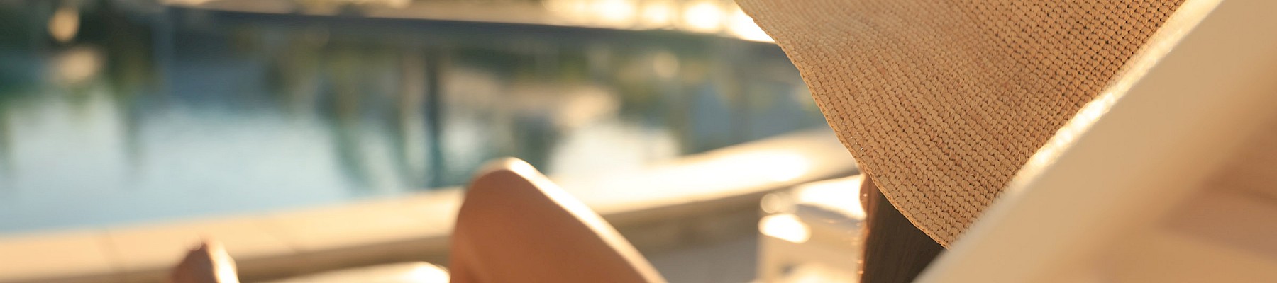A person wearing a straw hat lounging on a sun chair by a pool, enjoying the warm sun and relaxing vibes.