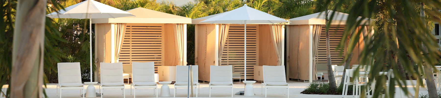 A resort pool with clear blue water, white lounge chairs, shaded cabanas, and palm trees framing a sunny, tropical scene.