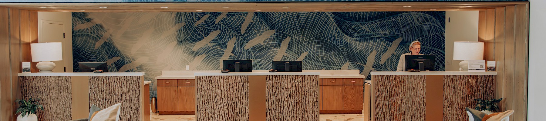 A modern hotel lobby with a front desk area, marble counters, tropical mural backdrop, warm wood ceiling, and comfortable seating in neutral tones.