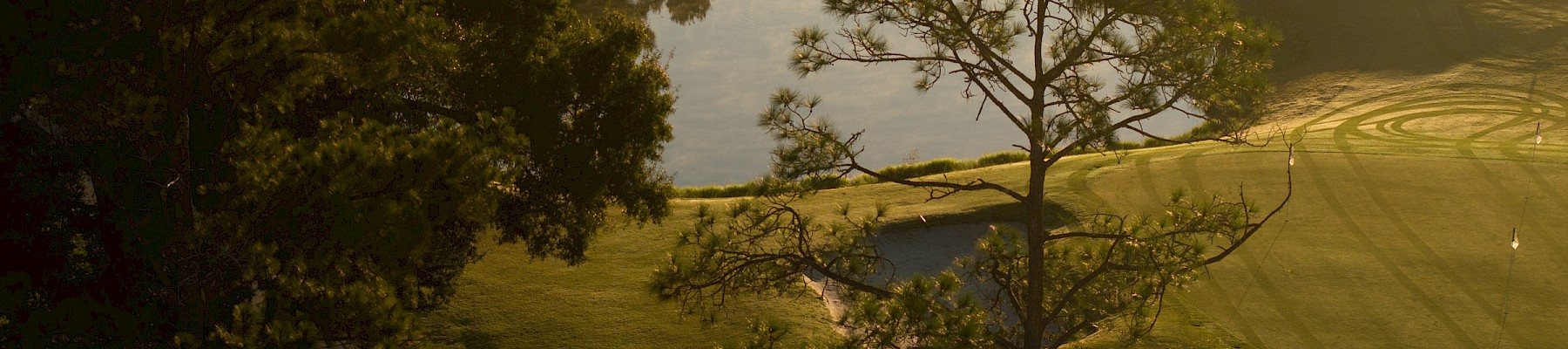 A serene golf course at dusk: winding path, lush trees, calm water reflecting the golden sky, and soft shadows across neatly mowed greens.