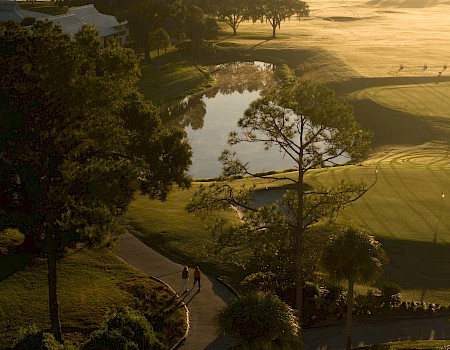 A serene golf course at dusk: winding path, lush trees, calm water reflecting the golden sky, and soft shadows across neatly mowed greens.