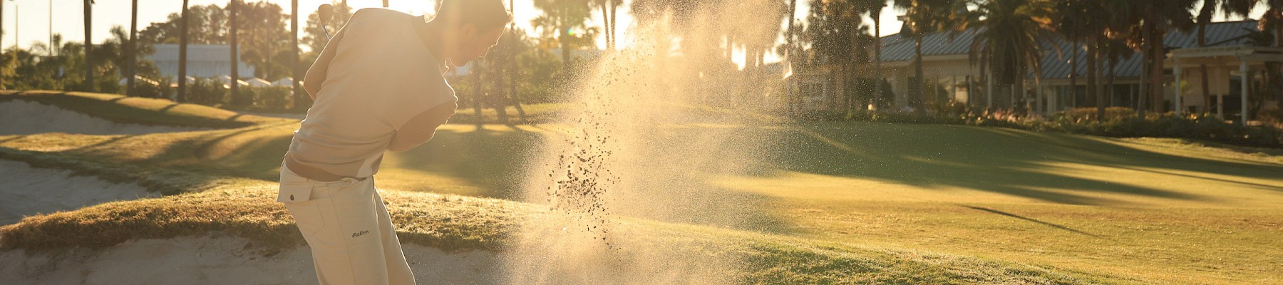 Golfer hits a sand shot from a bunker as the sun sets, sending up a spray of sand with palm trees lining the background.