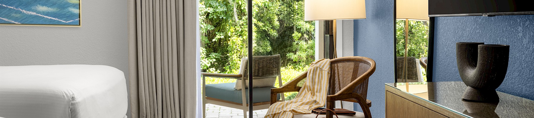 A modern hotel room with a sliding glass door to a balcony, a white bed, patterned carpet, woven chair, and a wooden dresser with a lamp, ending with a period.