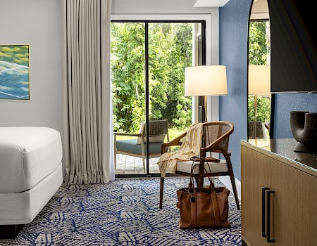 A modern hotel room with a sliding glass door to a balcony, a white bed, patterned carpet, woven chair, and a wooden dresser with a lamp, ending with a period.