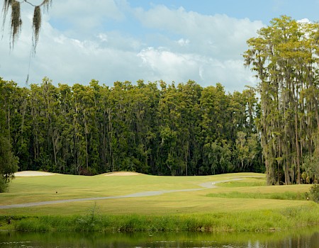 A peaceful golf course beside a small lake, framed by trees under a blue sky.
