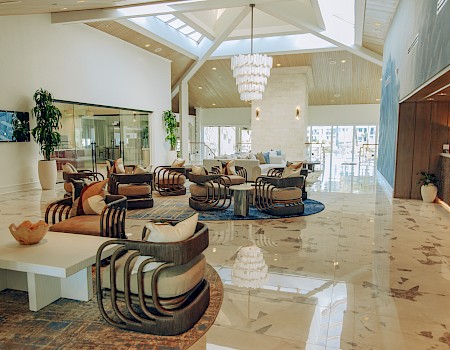 A bright hotel lobby with polished marble floors, modern armchairs, a chandelier, several plants, a reception area, and a seating lounge near large windows.