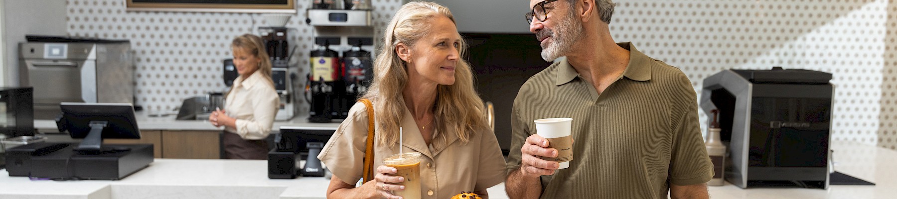 A stylish couple chats with coffee and pastries in a bright cafe, chatting cheerfully as they stand by the counter with warm lighting.