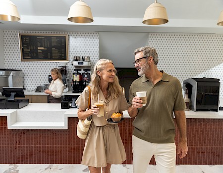 A stylish couple chats with coffee and pastries in a bright cafe, chatting cheerfully as they stand by the counter with warm lighting.