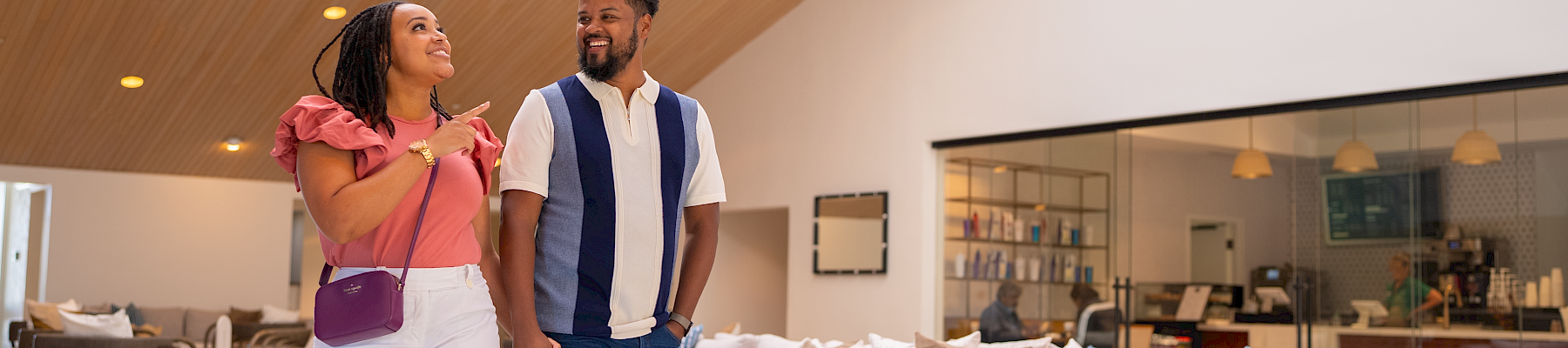A stylish couple strolling through a bright, modern hotel lobby with a chandelier, comfy seating, and warm wooden ceiling, smiling at each other.