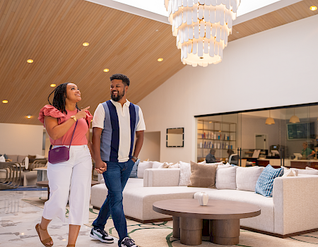 A stylish couple strolling through a bright, modern hotel lobby with a chandelier, comfy seating, and warm wooden ceiling, smiling at each other.