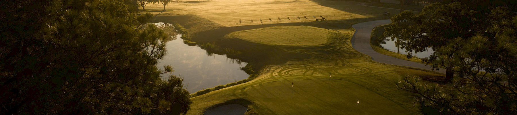 An aerial view of a golf course at sunset, with green fairways, sand bunkers, and trees framing the scene as the sun lights the horizon.