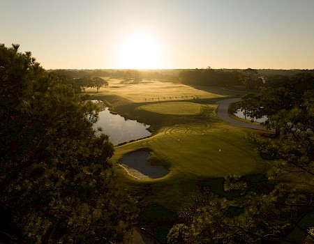 An aerial view of a golf course at sunset, with green fairways, sand bunkers, and trees framing the scene as the sun lights the horizon.