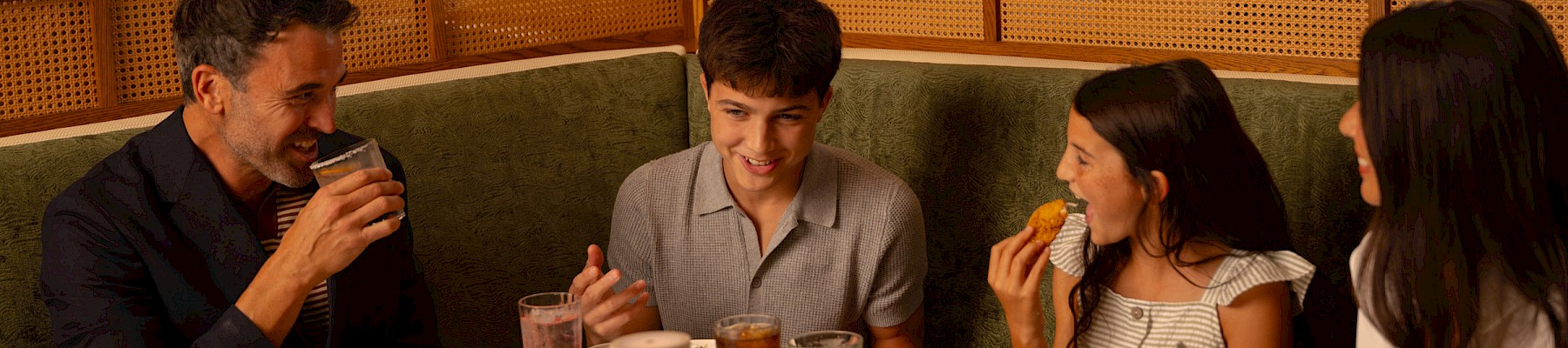 A family dinner at a cozy restaurant, sharing plates and drinks around a white table with warm lighting.