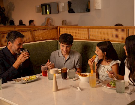 A family dinner at a cozy restaurant, sharing plates and drinks around a white table with warm lighting.