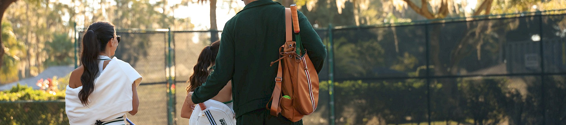 Three people walk off a tennis court together after a game, chatting as they carry rackets and bags, sunlight filtering through trees.