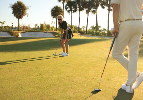 Two people playing golf on a sunny course; one putting, the other standing with a club, palm trees in the background.