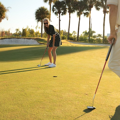 Two people playing golf on a sunny course; one putting, the other standing with a club, palm trees in the background.
