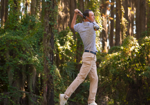 A man in light clothing swings a golf club on a sunny course, with tall trees in the background, mid-swing and focused. End the sentence.