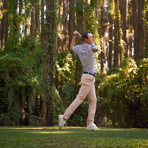 A man in light clothing swings a golf club on a sunny course, with tall trees in the background, mid-swing and focused. End the sentence.