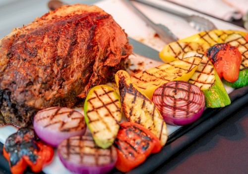 A charred grilled meat with vibrant orange-red crust sits beside colorful grilled vegetables—zucchini, onions, and peppers—on a sleek black platter.