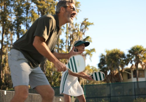 Two people playing beach tennis, paddle racket in hand, near a fence with palm trees in the background, smiling and ready to hit the ball.