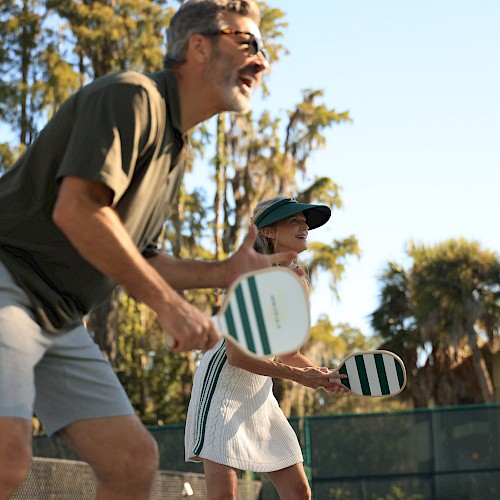 Two people playing beach tennis, paddle racket in hand, near a fence with palm trees in the background, smiling and ready to hit the ball.