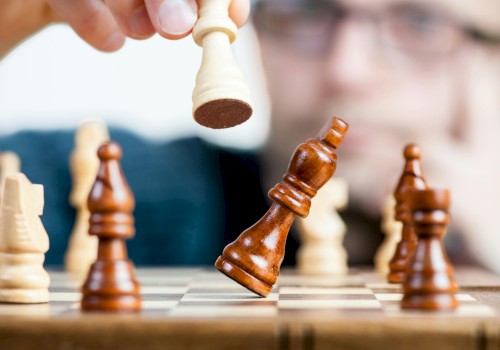 A person moves a white chess king, capturing a brown piece on a chessboard as others stand ready, focus on the game and strategy.