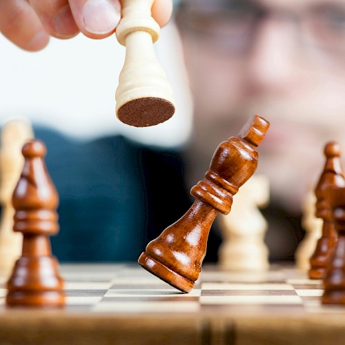 A person moves a white chess king, capturing a brown piece on a chessboard as others stand ready, focus on the game and strategy.