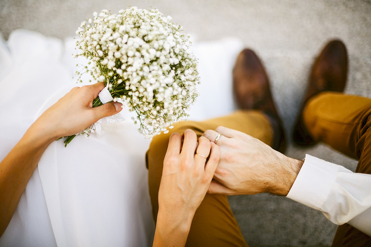 A person holds a small bouquet while another rests a hand on their knee. They both wear formal attire, facing each other.