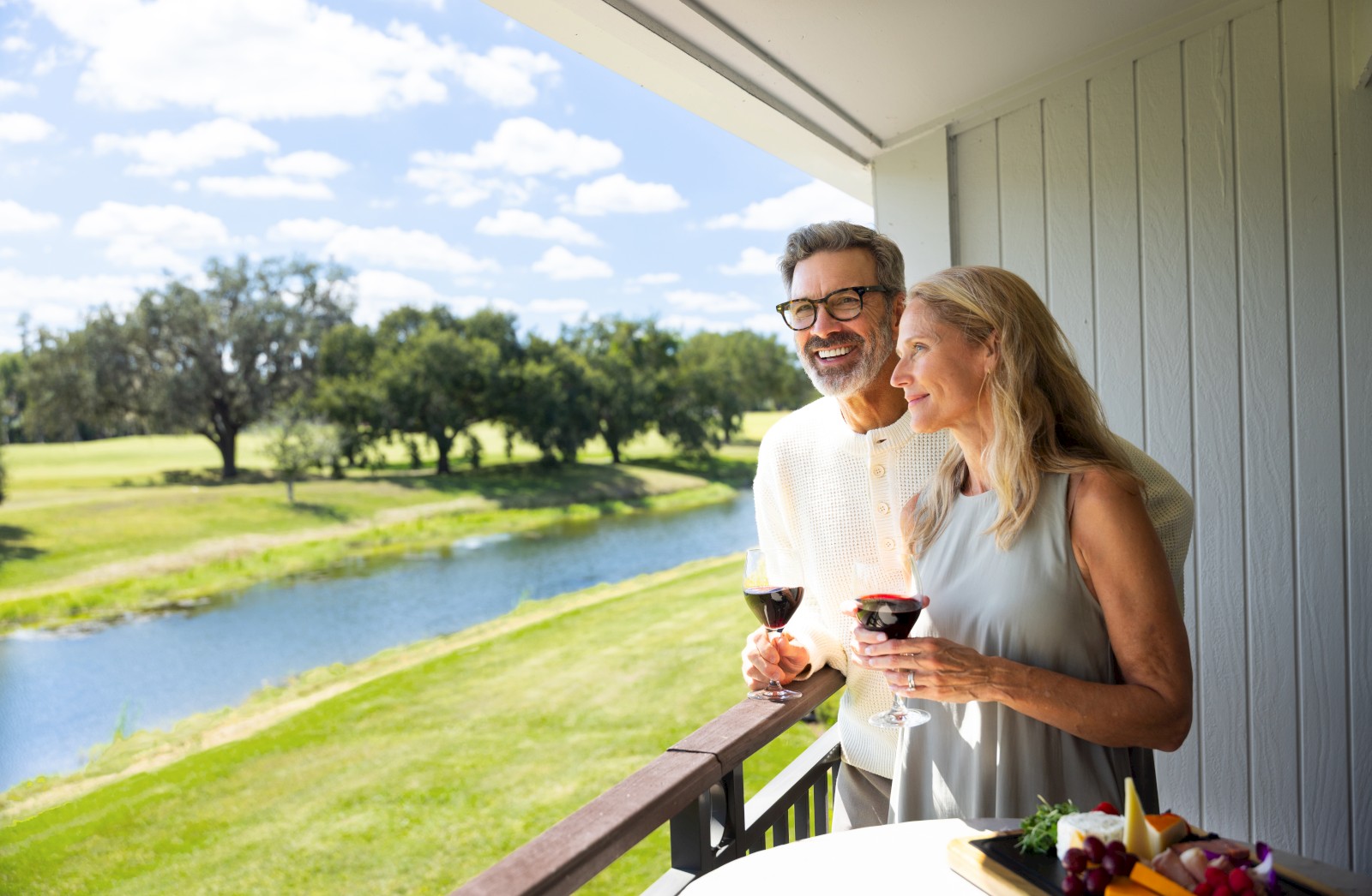 Two adults on a balcony overlooking a sunny golf course/lake, enjoying wine and a platter of food together, smiling at the scenery.