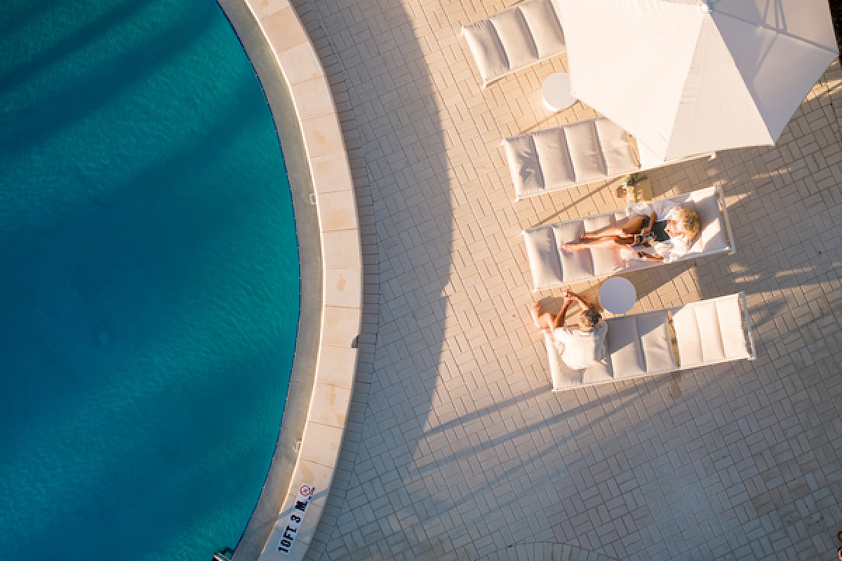 People relaxing on white loungers and umbrellas beside a curved blue pool at a sunny resort, enjoying a leisure day.