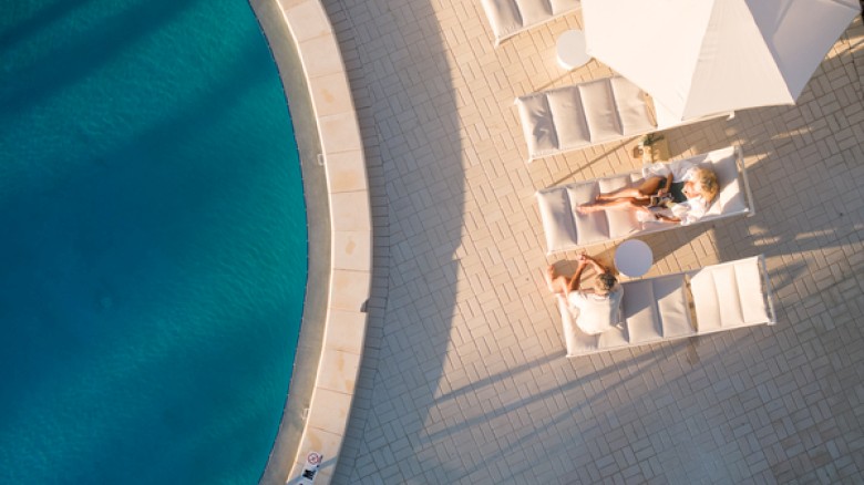 People relaxing on white loungers and umbrellas beside a curved blue pool at a sunny resort, enjoying a leisure day.
