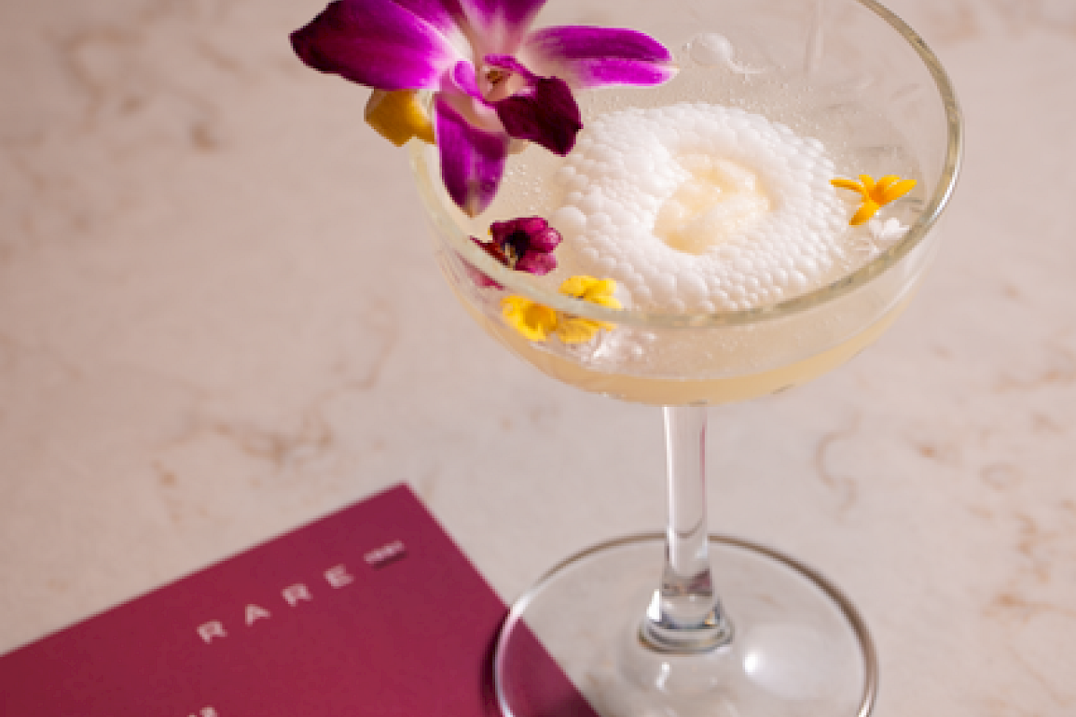 A stylish cocktail in a coupe glass with an orchid garnish, a foamy top, and a maroon menu card nearby on a light marble surface.