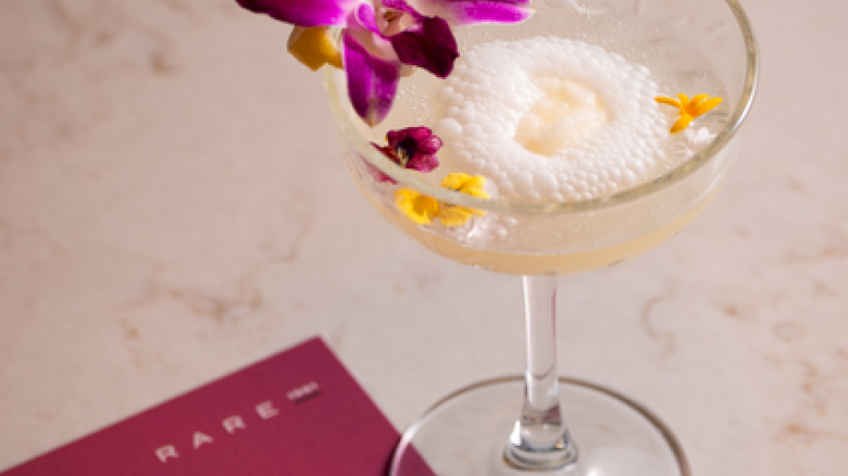A stylish cocktail in a coupe glass with an orchid garnish, a foamy top, and a maroon menu card nearby on a light marble surface.