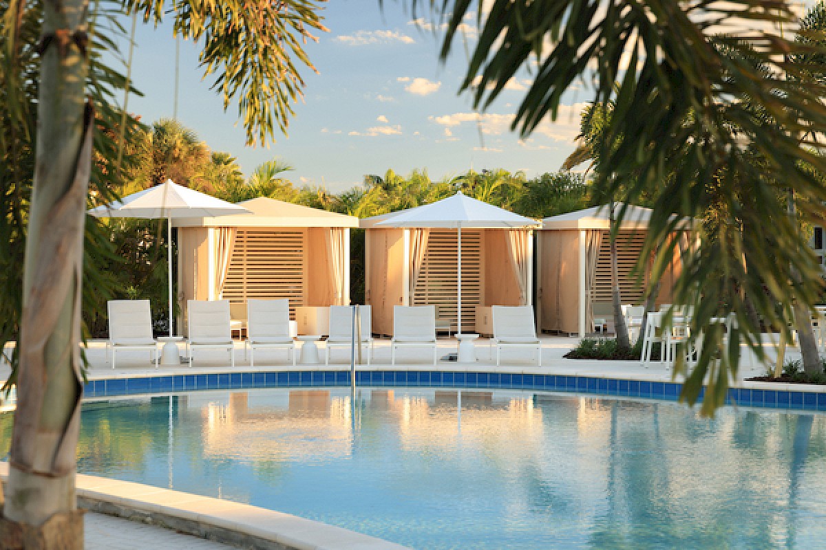 A resort pool with clear blue water, white lounge chairs, and shaded cabanas under palm trees by a sunny, tropical setting.