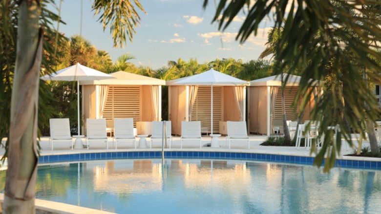 A resort pool with clear blue water, white lounge chairs, and shaded cabanas under palm trees by a sunny, tropical setting.