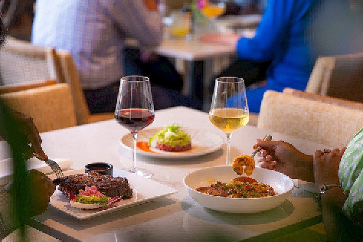 A group enjoys a meal: two people share red and white wine, a plate with steak, a salad, a bowl of pasta, and a small fried appetizer on a table.