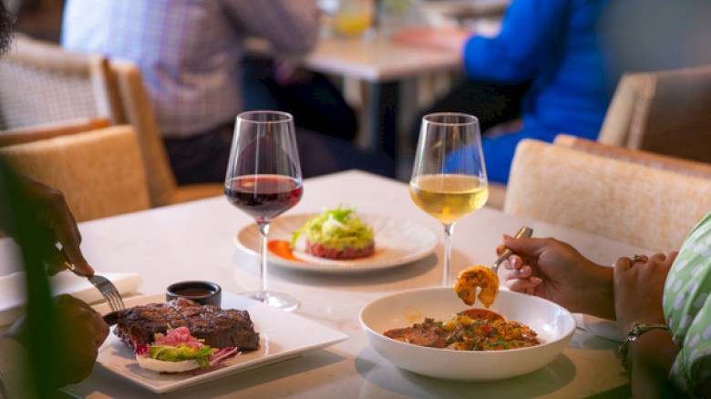 A group enjoys a meal: two people share red and white wine, a plate with steak, a salad, a bowl of pasta, and a small fried appetizer on a table.