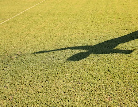 Shadow of a person or figure on a grassy field, long stretch of shade near the edge of a line, bright sunlight illuminating the grass.