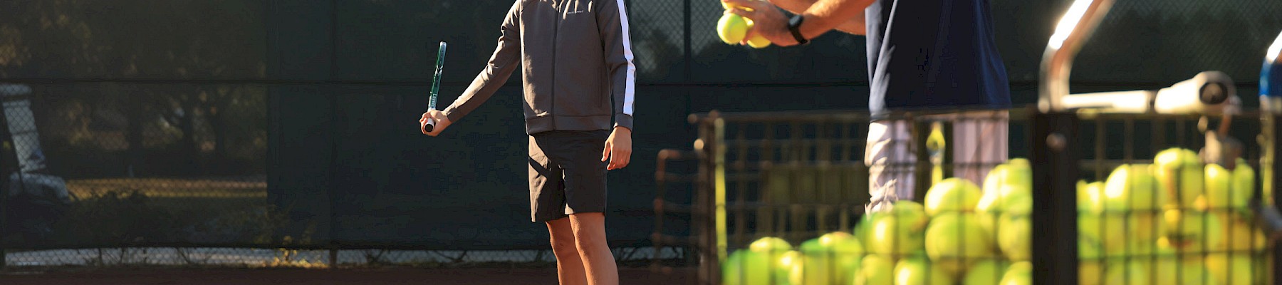 Two boys on a tennis court with a rack of tennis balls; one holds a racket while the other tosses a ball, ready to practice drills.