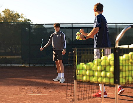 Two boys on a tennis court with a rack of tennis balls; one holds a racket while the other tosses a ball, ready to practice drills.