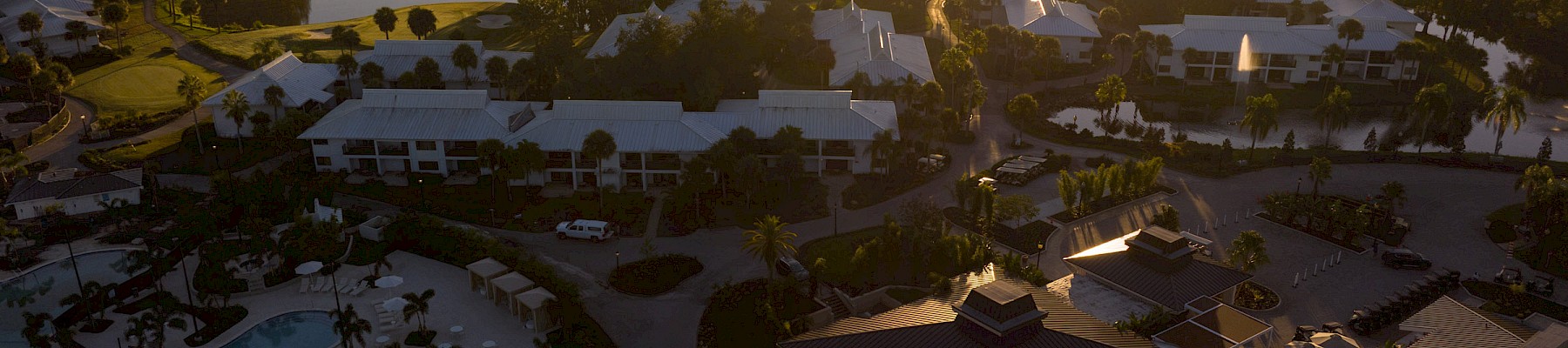 An aerial view of a resort complex at sunset, with pools, palm trees, and low buildings spread across a sprawling campus along a sunlit horizon.