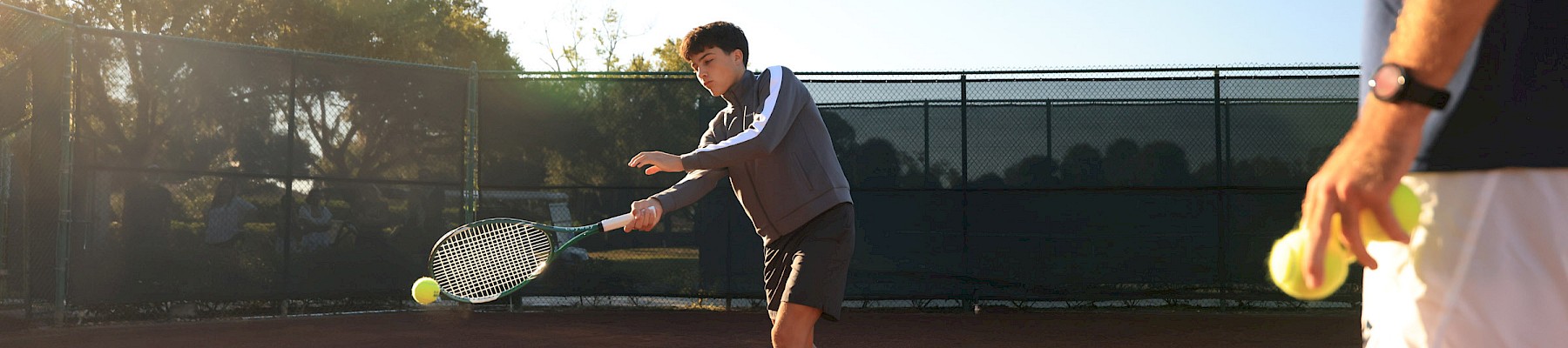 Two people on a tennis court: one prepares a forehand shot as the ball sails toward him, sunlight glints off the court and fence, tense practice.