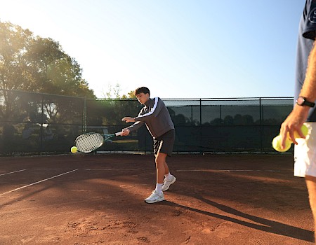 Two people on a tennis court: one prepares a forehand shot as the ball sails toward him, sunlight glints off the court and fence, tense practice.