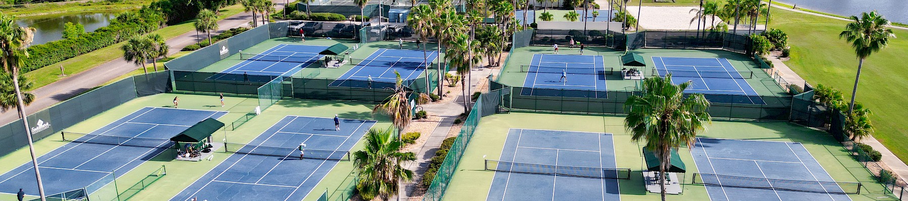 Aerial view of a tennis facility with multiple blue courts, green fencing, palm trees, and surrounding greenery, under sunny skies.