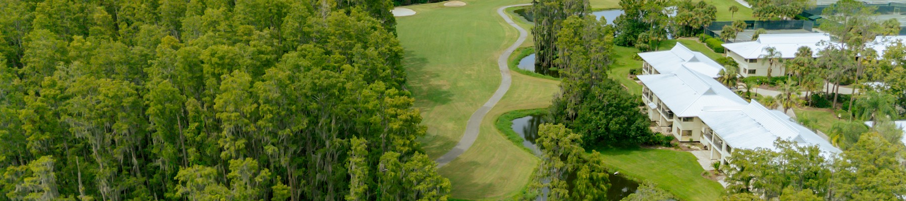 Lush green golf course nestled by a dense tree line, with a pond, winding cart paths, and white buildings along the edge under a partly cloudy sky.