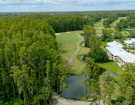 Lush green golf course nestled by a dense tree line, with a pond, winding cart paths, and white buildings along the edge under a partly cloudy sky.