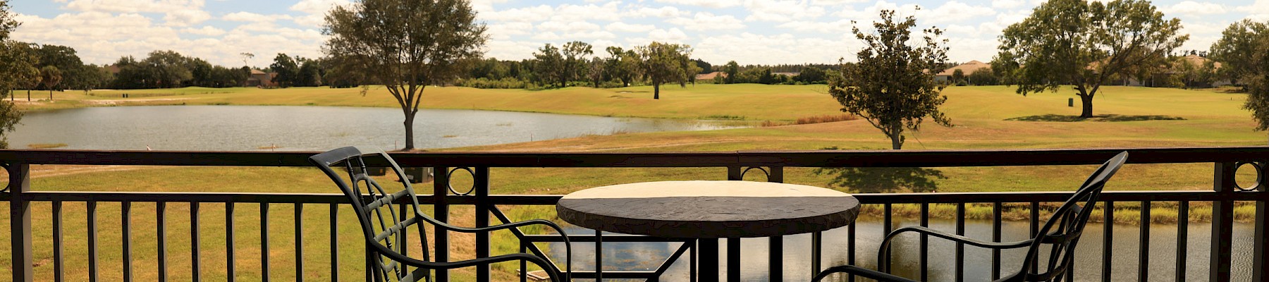 A sunny balcony view overlooks a grassy field with scattered trees, a round table and two chairs framed by a black railing.